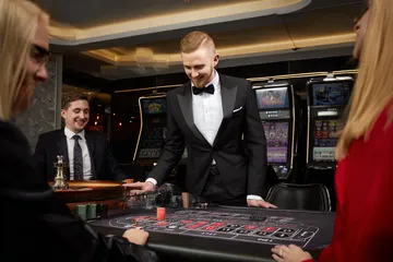 TAKSIM BET Slotlar — Binlerce tema ve jackpot sizi bekliyor Parlayan yedili sembolleri gösteren slot makinelerinin yanında şık bir kadın, TAKSIM BET’teki geniş slot çeşitliliğini gösterir.